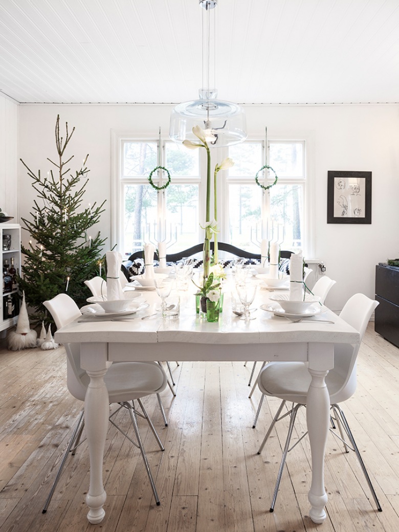 Typowo skandynawska dekoracja świątecznej jadalni, w której dominuje biel oraz zielone naturalne akcenty. Skromna zastawa na stole i choinka w swojej surowej postaci przełamują jasną kolorystykę wnętrza i wnoszą do niego delikatny gwiazdkowy...