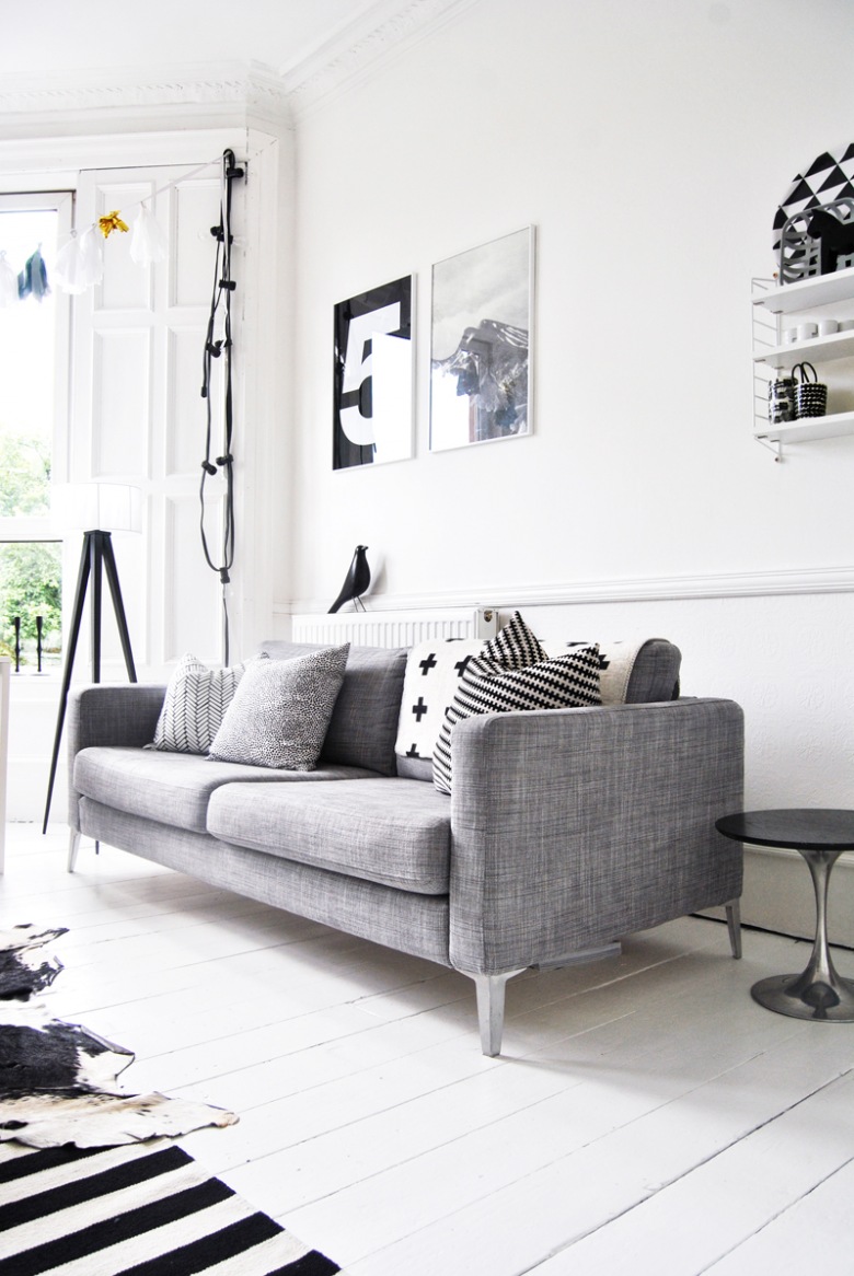 Białe sztukaterie na ścianach, drewniane okiennice i zarówki na kablu w salonie z szarą sofą i graficznymi dekoracjami (24889)
