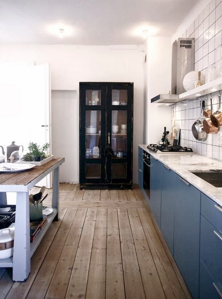 Niebieskie fronty szafek i drewniana podłoga  w kuchni z  czarną szafą tworzą wyśmienitą aranżację.Blat wyspy nawiązuje do wyjątkowej...