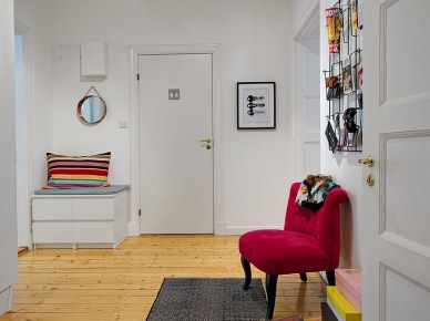 Drewniane deski na podłodze,różowy fotelik,biała komoda na buty z szarym siedziskiem i kolorową poduszką w paski,grafika czarno-biała,okragłe lustro na scianie w przedpokoju (25769)