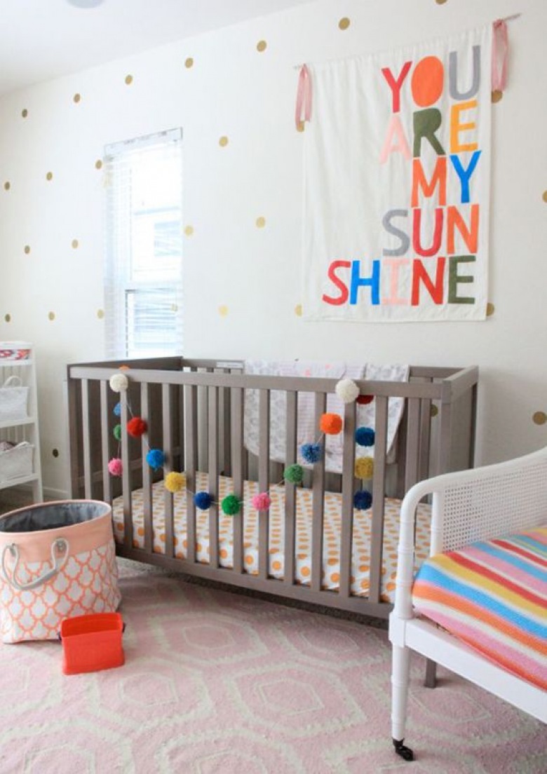 Kolorowe dekoracje mocno urozmaicają wystrój pokoiku dziecięcego. Proste łóżeczko nawiązuje do stylu skandynawskiego.