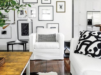 Galeria biało-czarnej fotogreafi i grafik na białej ścianie w salonie z białymi fotelami i sofą i rustykalnym stołem z drewnianym blatem (26785)