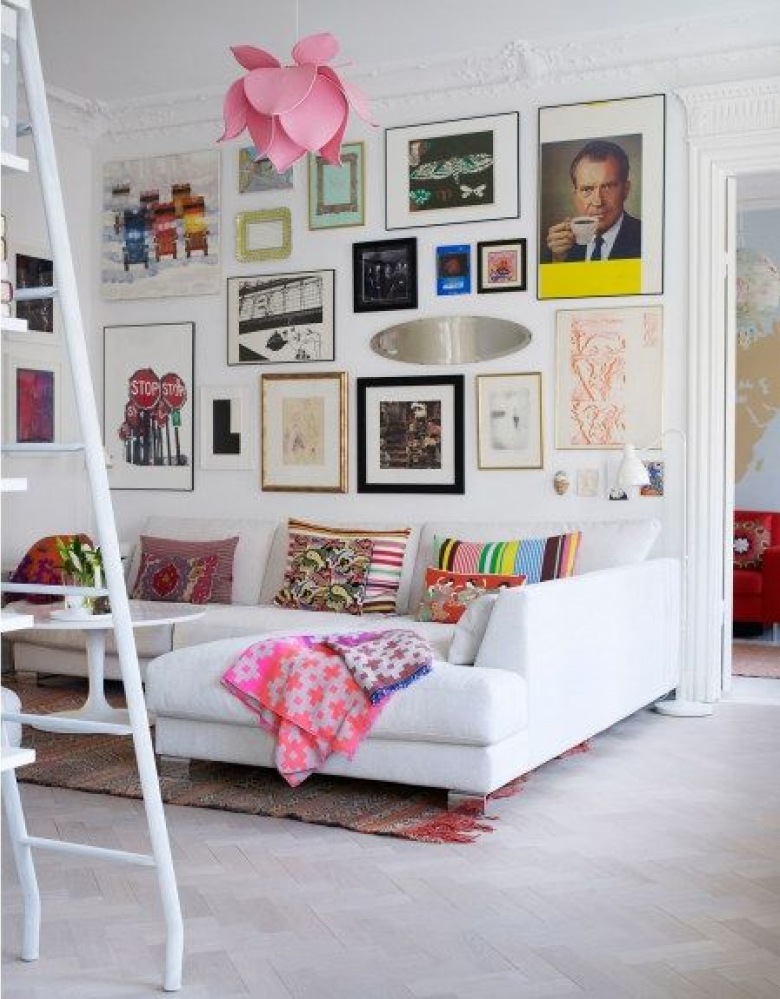 Pomysł na kolorową galerię na białej ścianie z grafik i plakatów i róznobarwne poduszki na sofie w salonie (28057)