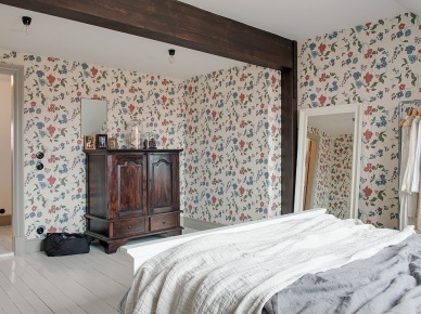 Drewniane ciemnobrązowe belki w sypialni,kwiatowa tapeta ścienna w niebiesko-czerwonym kolorze,retro komoda bieliźniarka przy ścianie,duże białe lustro stojące (25942)