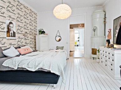 Drukowana tapeta z napisami,białe deski na podłodze,biała komoda szufladnik i czarne łóżko tapicerowane w skandynawskiej sypialni (28413)