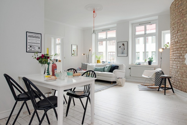 Biały stół z czarnymi krzesłami w otwartej przestrzeni mieszkania z salonem z białą podłogą i ścianą z czerwonej postarzanej cegły (26463)