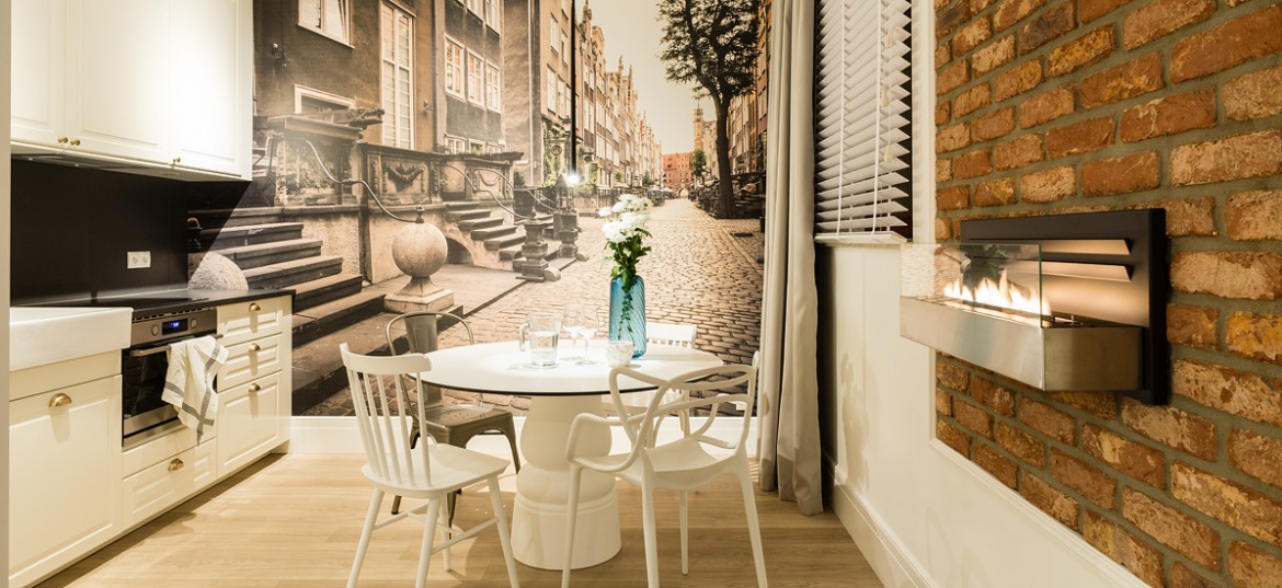 Fototapeta z ulicą Mariacką w Gdańsku stanowi tu tło dla jadalni i kuchni. Pomieszczenie utrzymane w kolorach brązowym,...