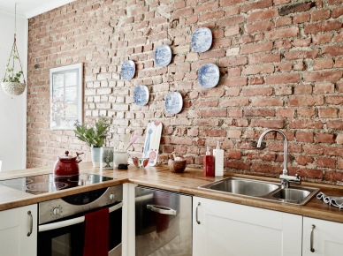Ściana z czerwonej cegły,ozdobne biało-niebieskie talerze na ścianie  i biała kuchnia skandynawska z drewnianymi blatami (27036)