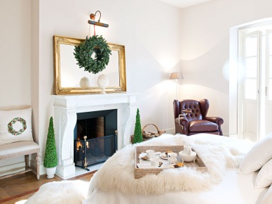 Biała sypialnia wiele zyskuje dzięki drobnym, ale wyrazistym świątecznym dodatkom. W głównej mierze wybrano zielone naturalne gałązki, układając kompozycję w wieniec, albo dekoracyjne drzewka. Miękkie oświetlenie dodaje ciepła i przytulności do wnętrza, podobnie, jak kominek naprzeciw...